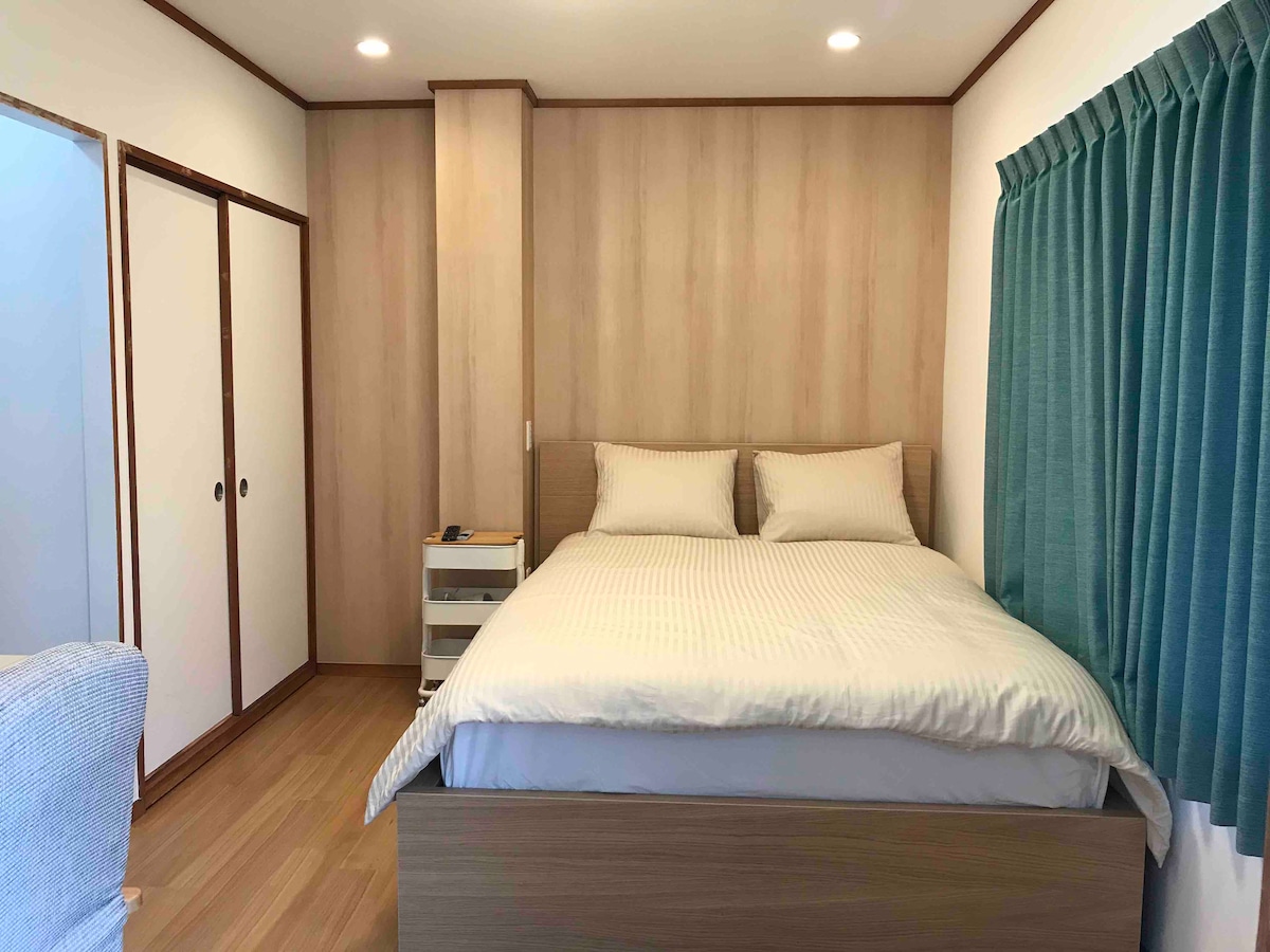 A well-arranged sleeping area features a spacious bed with two pillows. A built-in closet is visible to the left, alongside a nightstand. Light-colored wood panels cover the walls, and a teal curtain frames the window, allowing natural light to enter the room.