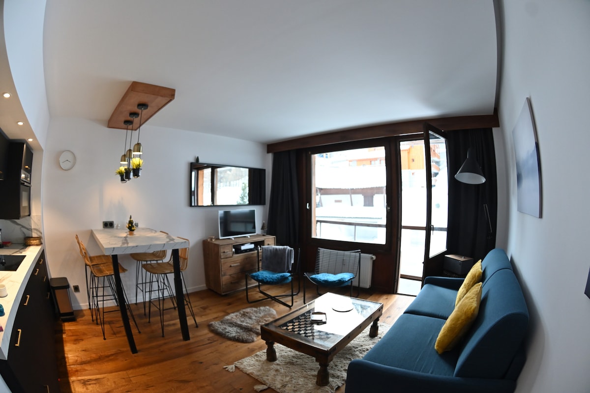 A renovated studio is showcased, featuring a cozy seating area with a blue sofa and a wooden coffee table. A dining table with modern chairs is present, along with a television mounted on the wall. Large windows provide natural light and views of the surrounding area.