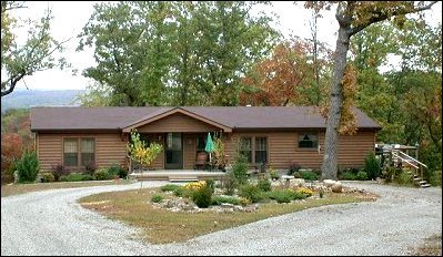 A log style home is set amidst a landscaped yard, surrounded by greenery. A circular gravel driveway leads to the entrance, accentuated by planters and an umbrella-covered seating area. Large windows provide views of the outdoors, with subtle autumn colors highlighted by the trees.