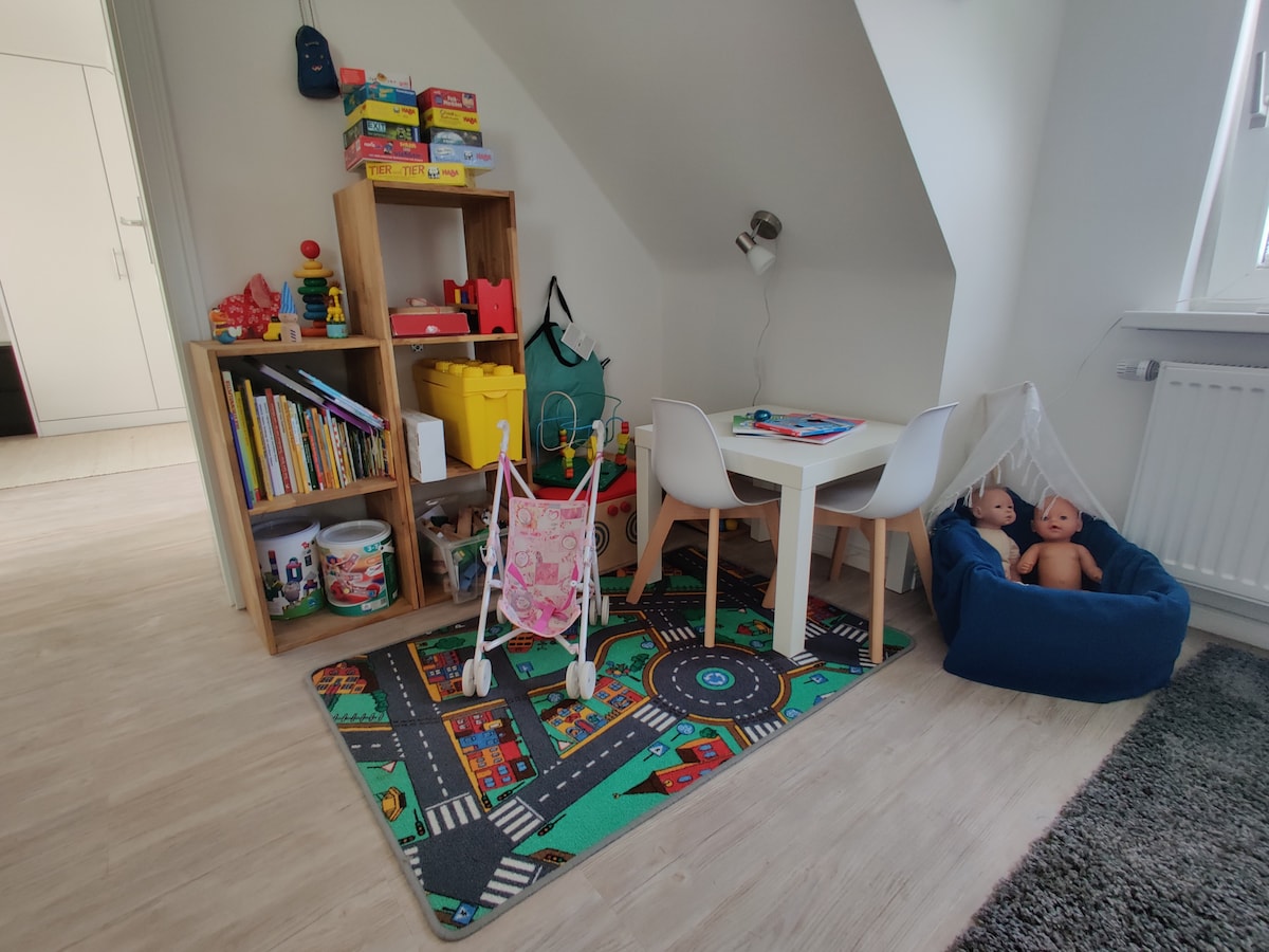 A children's play area is highlighted, featuring a colorful rug with road patterns. A small table with white chairs sits beside a bookshelf filled with various toys and books. Soft toys are displayed in a cozy nook, creating an inviting space for play.