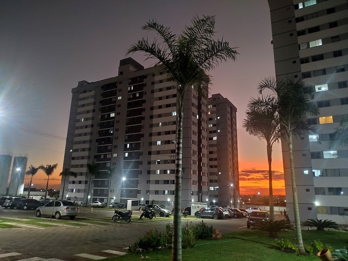 The exterior of the apartment building is highlighted against a vibrant sunset, with large windows reflecting the warm colors. Palm trees line the area, while parking spaces show multiple vehicles. The well-lit setting contributes to a welcoming evening atmosphere.