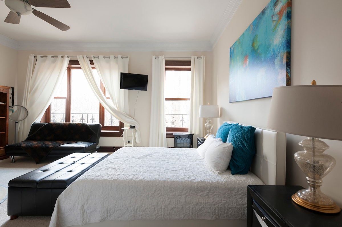 A spacious room is highlighted by large windows bringing in natural light, equipped with a comfortable bed featuring luxury linens. A sleek black leather sofa and a television complete the setting, while art decorates the walls, enhancing the room's character.