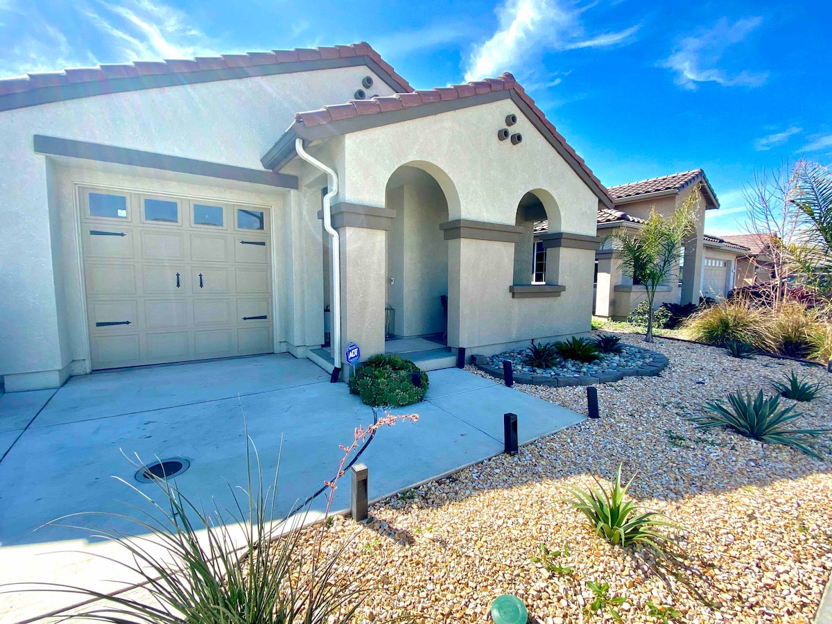 The exterior of the home is viewed, showcasing a single-story structure with a decorative entryway. A driveway leads to a spacious garage. Surrounding landscape features low-maintenance gravel and various shrubs, with palm trees gently swaying under a clear blue sky.