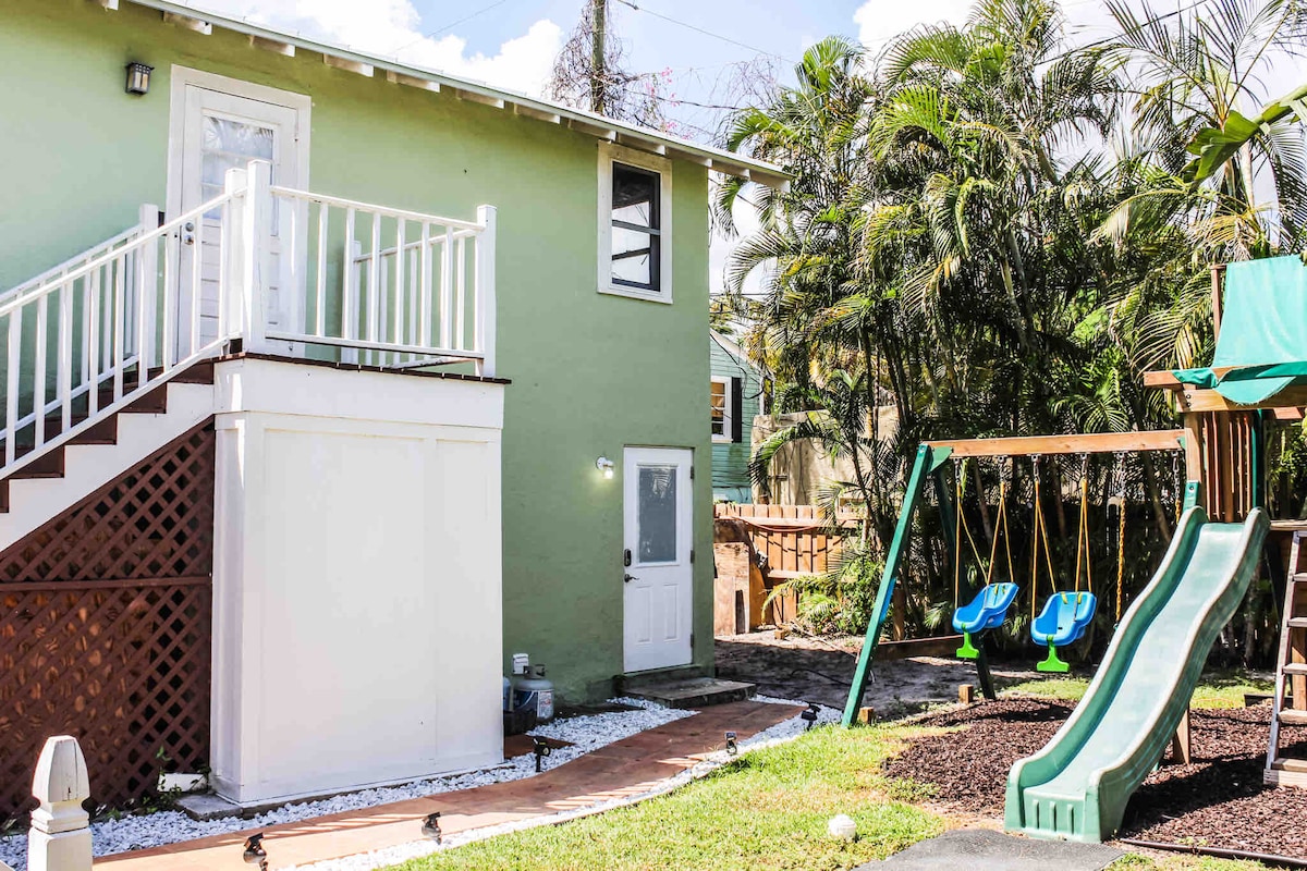 The exterior of the top-floor apartment features a green facade and a staircase leading to the entrance. A play area is visible, equipped with swings and a slide, surrounded by landscaped grass and palm trees, creating a welcoming outdoor space.
