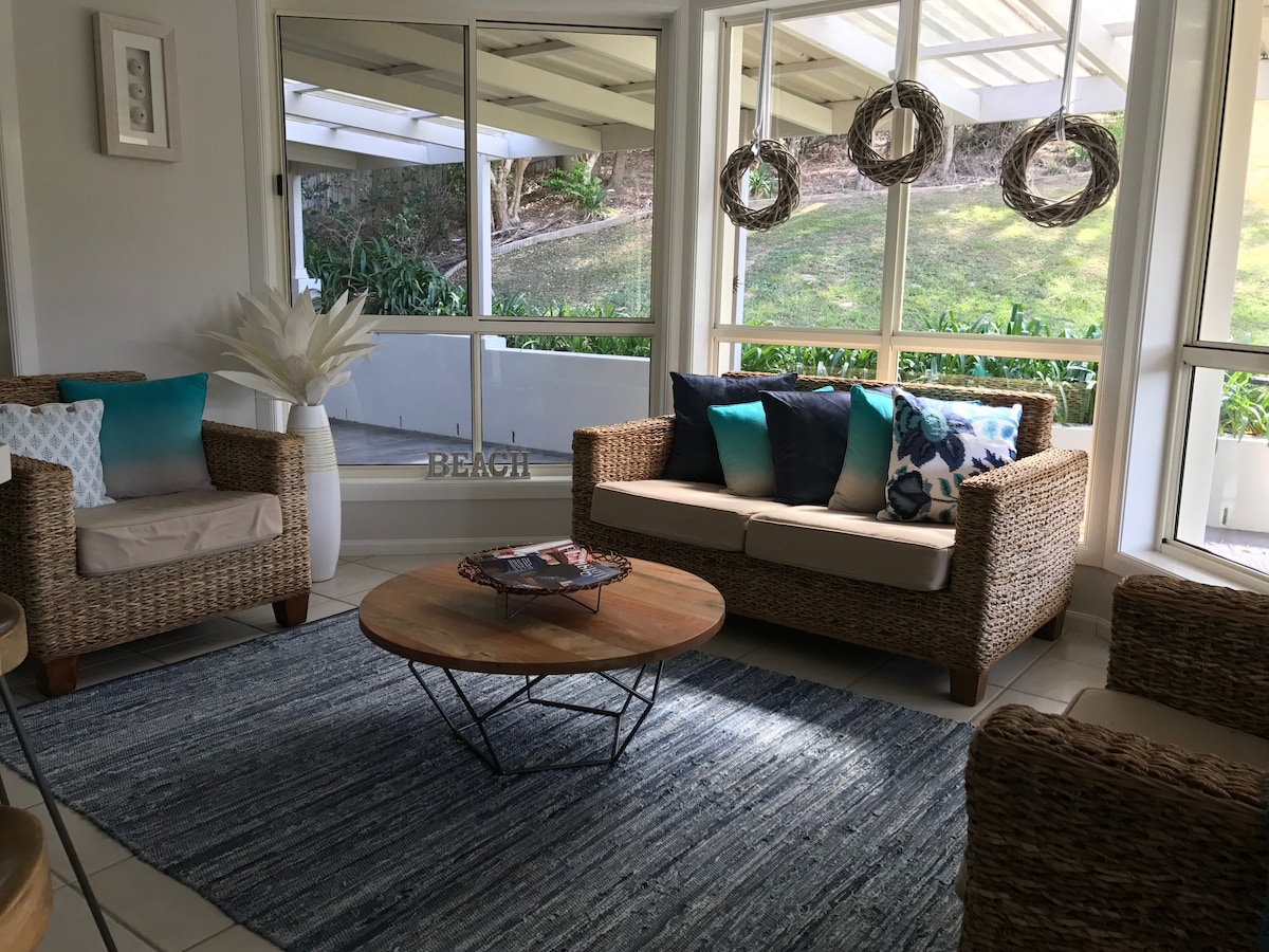 A sunlit living area features natural wicker chairs adorned with blue and white cushions. A round coffee table rests on a textured blue rug, complemented by light filtering through large windows overlooking a lush outdoor space.