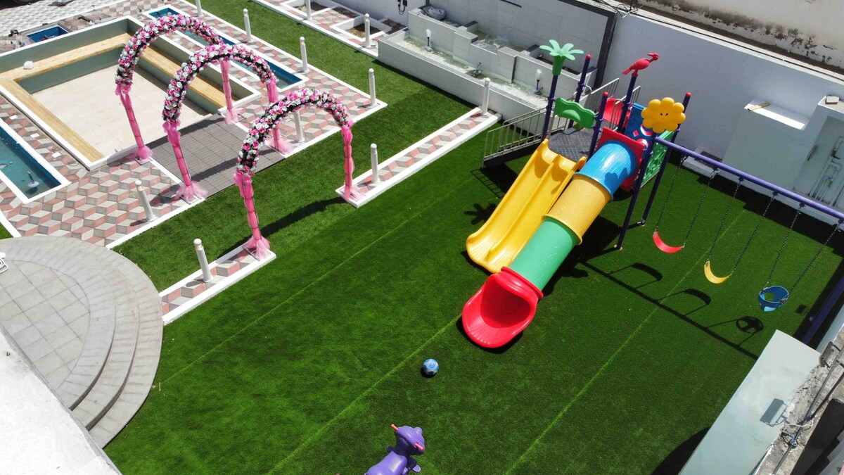 A spacious outdoor area features a vibrant playground with a colorful slide and swings, surrounded by well-maintained artificial grass. Two decorative arches are adorned with pink flowers, and a swimming pool is visible in the background, complementing the recreational space.
