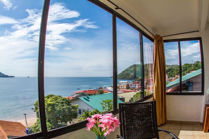 ห้องสว่างไสว มีเตียงควีนไซส์ไม้ เครื่องนอนสีฟ้า และหน้าต่างบานใหญ่พร้อมม่านบางๆ ที่มองเห็นวิวทะเลได้โดยตรง มีโต๊ะข้าง เก้าอี้ และมุมนั่งเล่นสบายๆ ให้ผ่อนคลาย ☀️