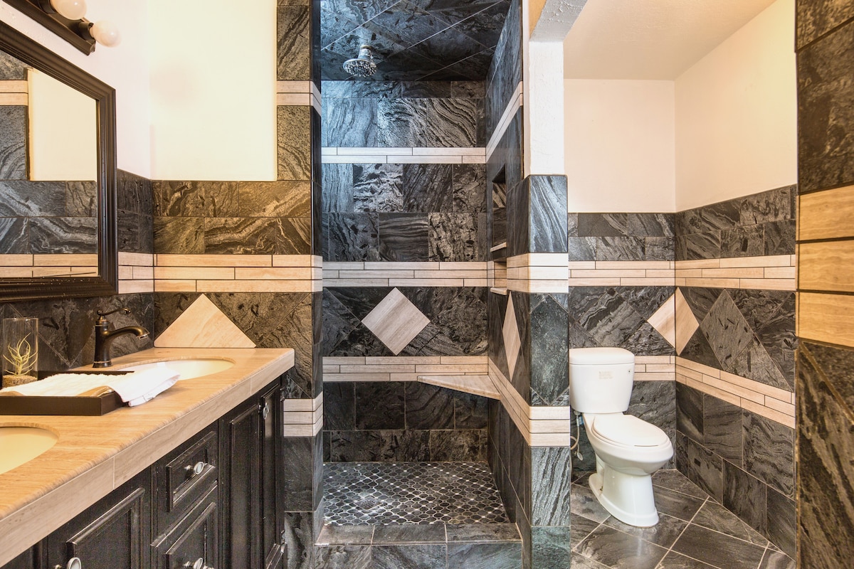 The bathroom features a dark stone design with contrasting light and dark tiles. A spacious walk-in shower is visible alongside a modern sink and vanity, and a toilet is positioned to the side. Natural light illuminates the room from a nearby source.