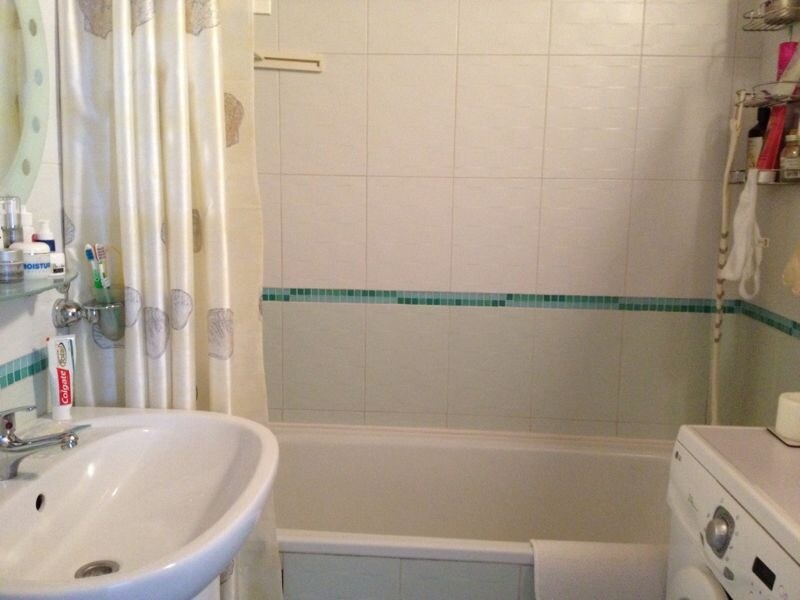 A compact bathroom features a washbasin with a mirror above, and a bathtub with a turquoise tile accent. A washing machine is positioned nearby, and a curtain adds privacy to the space. The overall decor is simple and functional, emphasizing cleanliness.