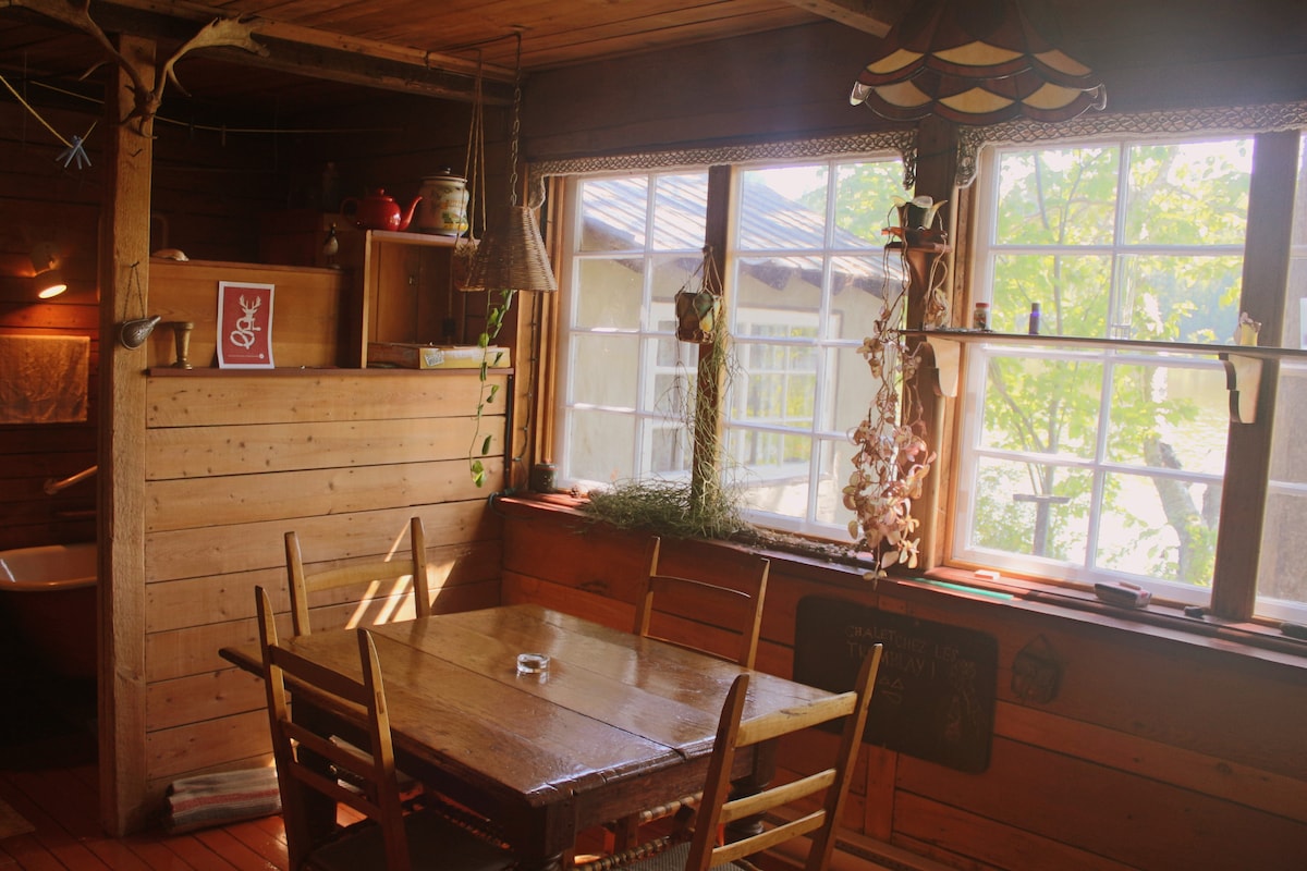 A wooden dining area features a simple table surrounded by several chairs. Sunlight filters through large windows, illuminating the space. Wooden walls and a few decorative elements enhance the cozy ambiance. A view of greenery is visible outside, adding to the serene setting.