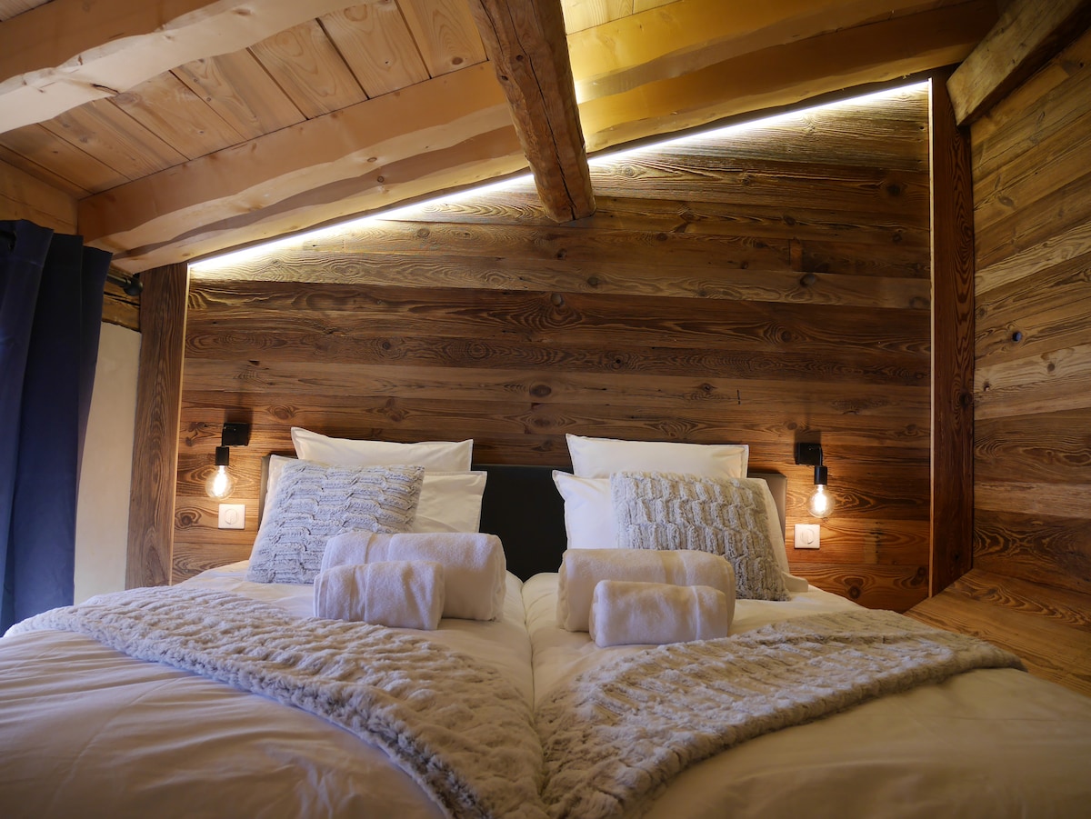 A warm sleeping area features two single beds, dressed in white linens and accented by soft, textured blankets. Wooden walls provide a rustic feel, illuminated by subtle lighting. Each bed is accompanied by a plush towel and a bedside reading light.