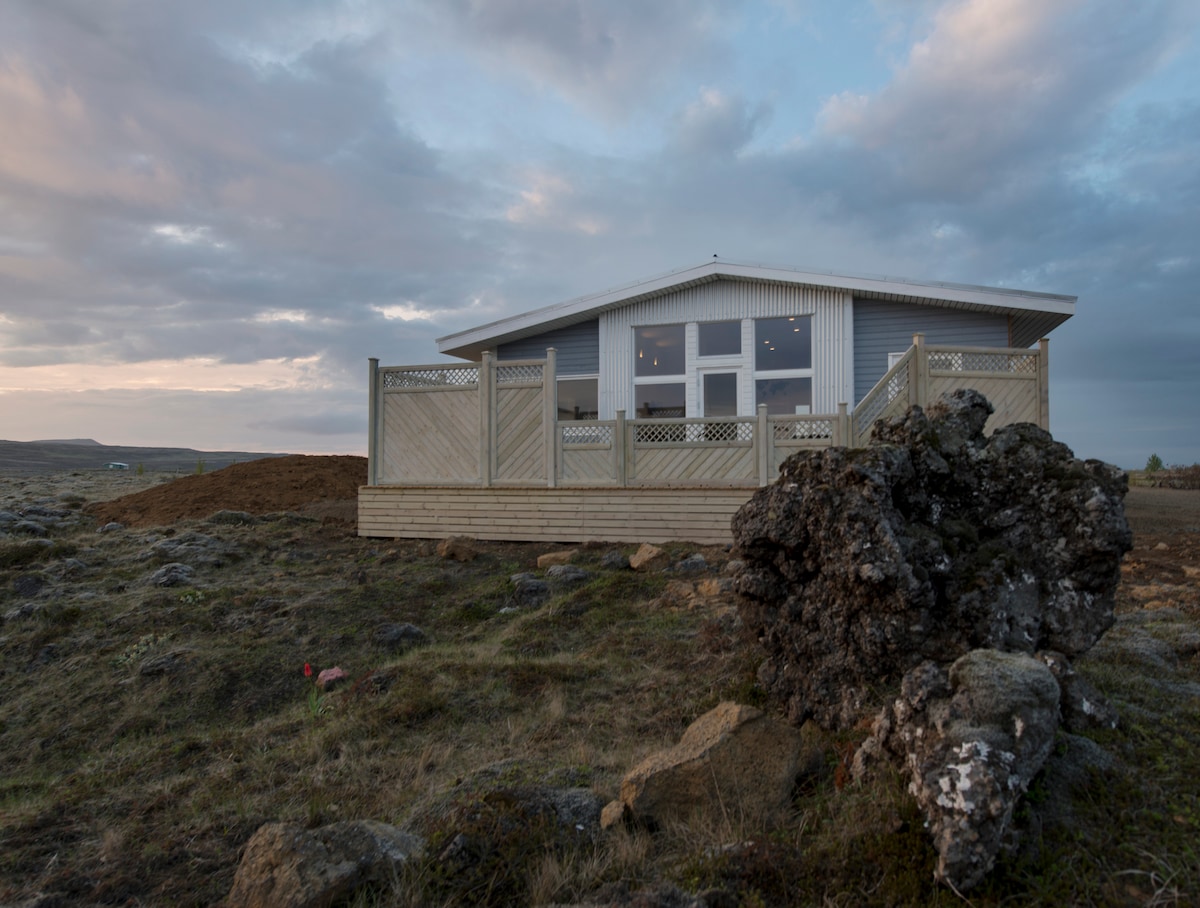 A modern home is showcased against a serene landscape, surrounded by rocky terrain and wild grass. The structure features a spacious wooden deck with railing, inviting outdoor seating to enjoy the surrounding scenery. Soft clouds drift across the sky, adding to the tranquil setting.