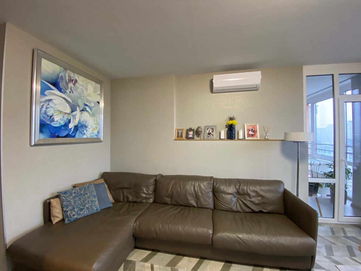A modern living area features a large sectional leather sofa, complemented by a decorative blue pillow. A floral artwork hangs above the sofa, and a shelf displays small framed photos and a decorative plant. Natural light enters from an adjacent glass door.