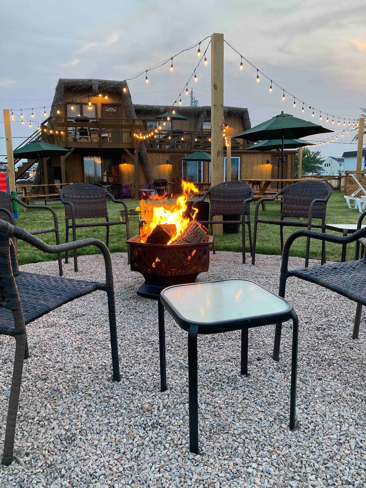 A gathering area features a fire pit surrounded by five wicker chairs arranged on gravel. Strung lights illuminate a wooden structure in the background, which includes a second floor. Green umbrellas provide shade over additional seating options nearby.