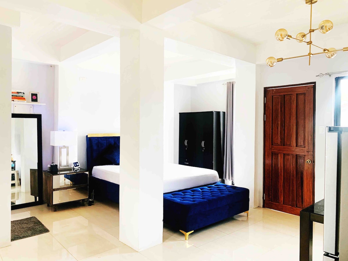 A modern studio features a spacious bed with a deep blue upholstered headboard. A stylish mirror and a small table with a lamp are positioned beside the bed. A dark cabinet contrasts against the light tiled flooring. An inviting natural light fills the room through large windows.