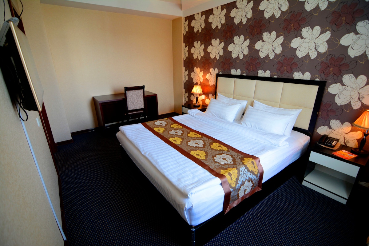 A cozy bedroom features a spacious bed with crisp white linens, adorned with a patterned throw blanket. Decorative wallpaper of large floral motifs lines the walls. A bedside lamp and a small desk with a chair provide additional furnishings in the room.