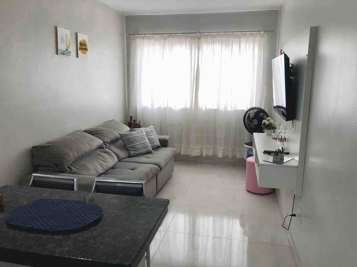 A living area features a comfortable gray sofa arranged near a wall-mounted television. Light filters through sheer curtains covering a large window, while a glass table with metal chairs occupies a corner of the space, creating a simple, functional layout.