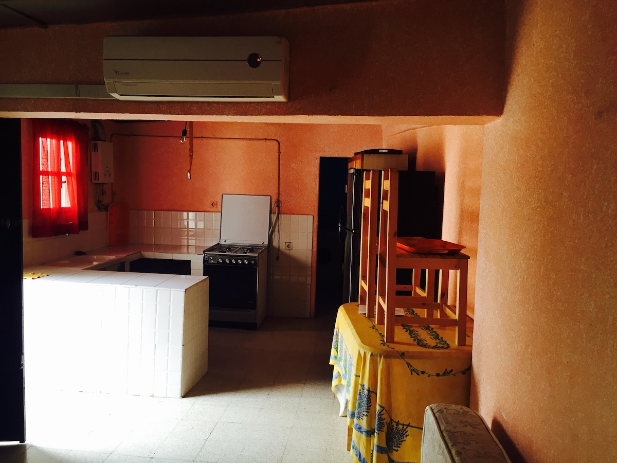 The kitchen area is defined by textured pink walls and features a large window draped with red curtains. A white countertop runs along one side, while a classic stove is visible in the corner. A table covered with a patterned cloth occupies the center, surrounded by wooden chairs.