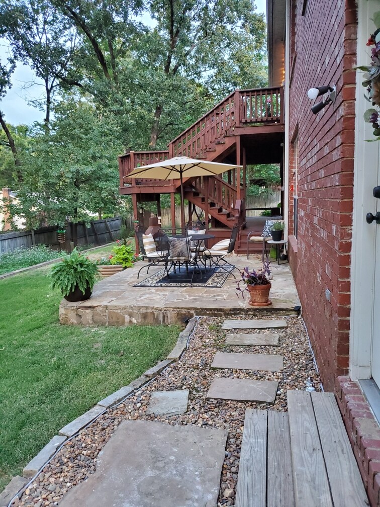 A private flagstone patio features a seating area with a round table and four chairs, surrounded by greenery. A large umbrella provides shade over the seating arrangement, with a pathway of stepping stones leading through a well-maintained yard. The patio is complemented by a wooden deck above.