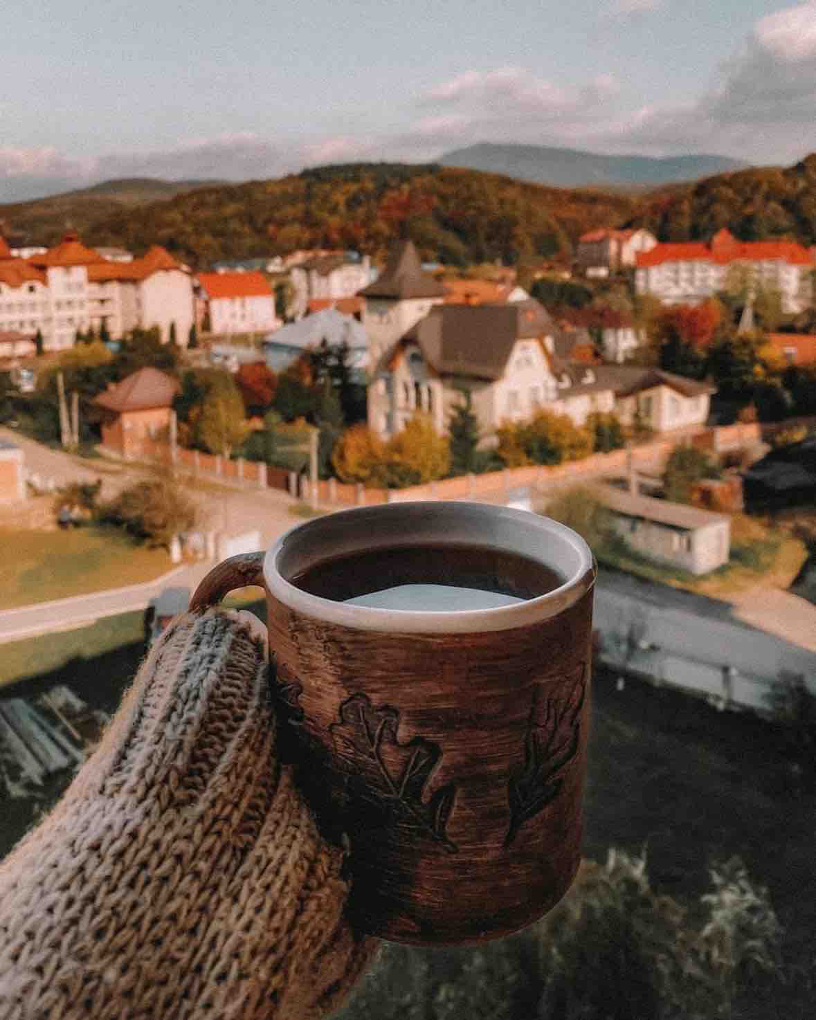 A hand holding a mug is seen in the foreground, with a scenic view of the village and surrounding mountains in the background. The landscape features a mix of residential buildings and greenery, highlighted by soft afternoon lighting.