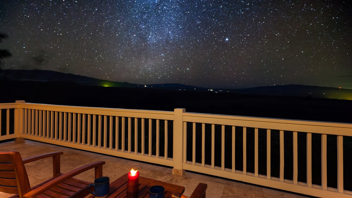 The private lanai overlooks a clear night sky filled with stars, offering expansive views of the landscape below. A lantern and two cups sit on a table next to rustic wooden chairs, inviting quiet moments under the stars.