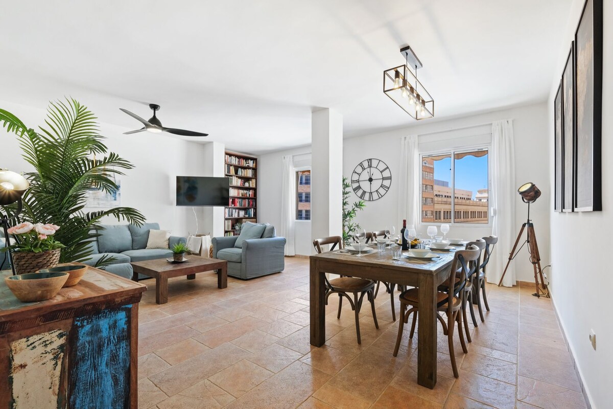 A spacious living area features a comfortable seating arrangement with gray sofas and a wooden dining table. A television is mounted on the wall, while large windows allow natural light to fill the room, offering views of the outdoors. Decorative plants add a touch of greenery.