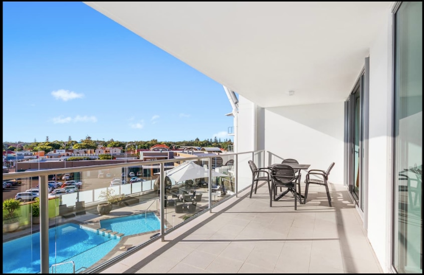 A spacious balcony features a glass railing overlooking the pool, with a dining set that includes four chairs and a round table. The bright blue sky and surrounding greenery create a pleasant outdoor setting.