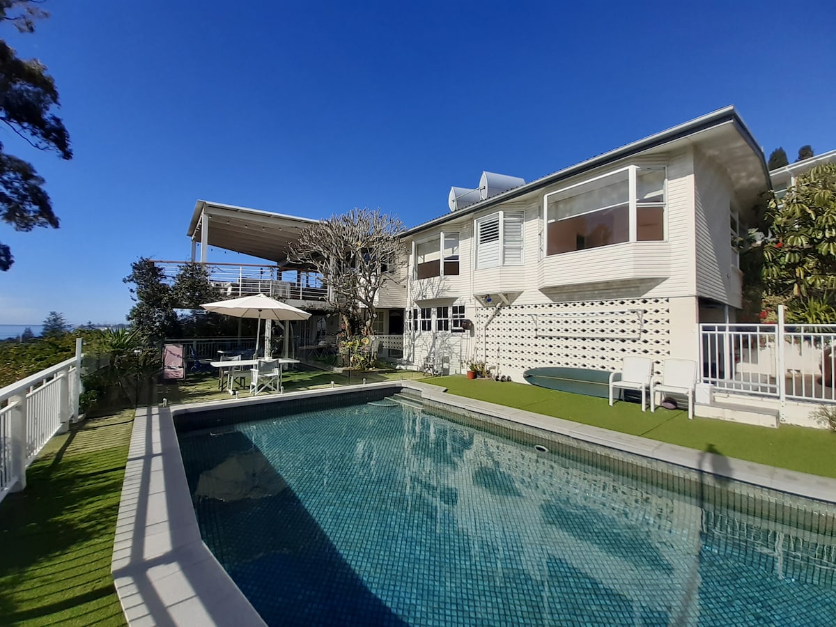 The exterior of a two-story house is captured, featuring large windows and a spacious outdoor area. A swimming pool is situated in front, surrounded by green landscaping. Sun loungers are arranged by the pool, while an umbrella provides shade. The sunlit blue sky enhances the overall scene.