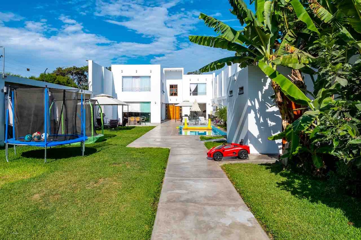 A well-maintained garden pathway leads to a modern two-story house. To the left, a trampoline is positioned on green grass, while a small play area with a pool is visible on the right. A toy car rests on the pathway, surrounded by tropical plants.