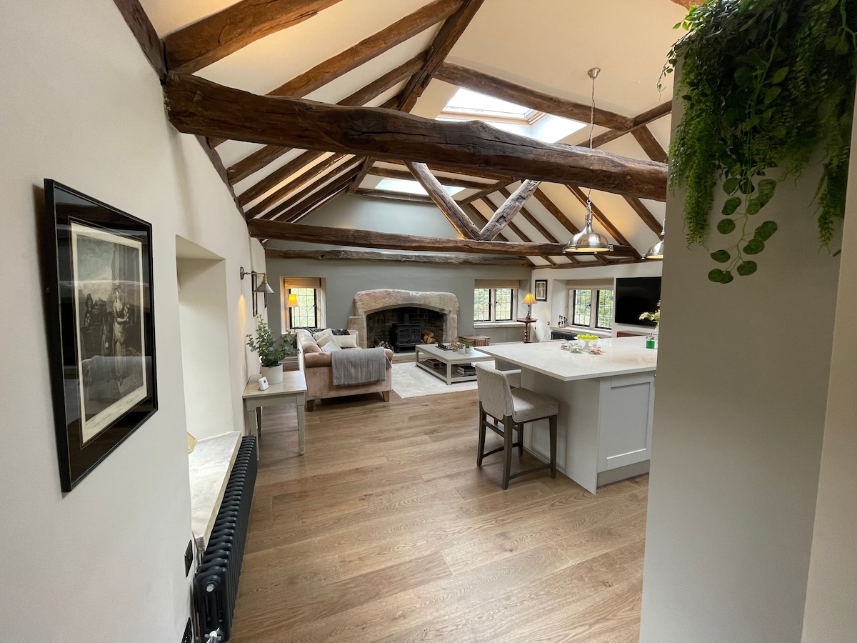 The open plan living area features exposed wooden beams and a light, airy atmosphere. A large sofa and coffee table are arranged around a central fireplace. A kitchen island with seating is visible, and natural light floods the space from several windows.