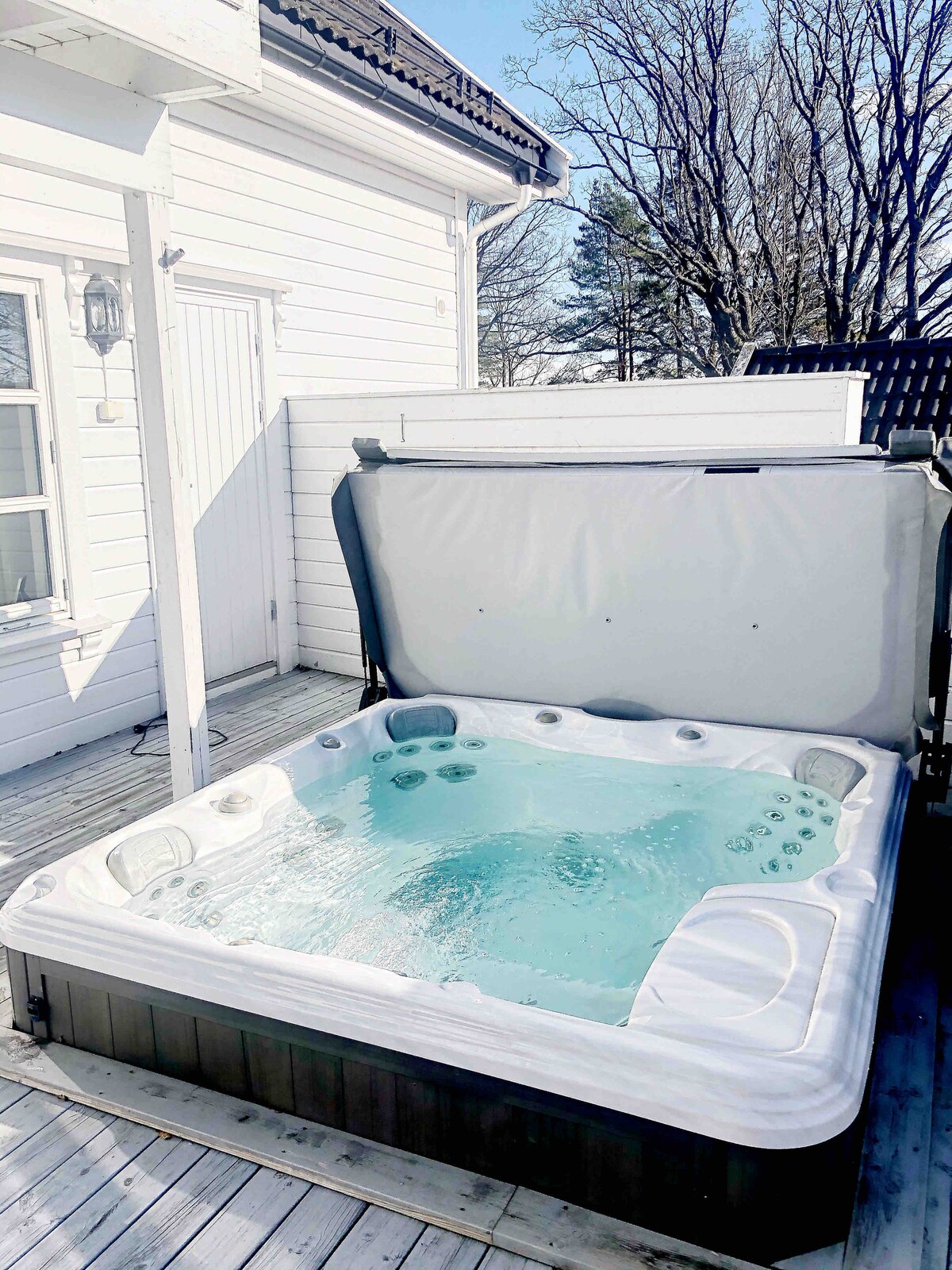 A Sundance Certa SPA is positioned on a wooden deck outside, featuring a spacious layout and sparkling water. The hot tub is covered with a liftable canopy, providing easy access for relaxation. Sunlight reflects off the water, creating an inviting atmosphere.