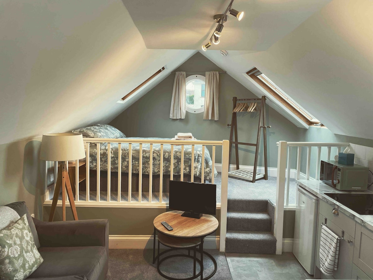 A mezzanine sleeping area is positioned under a sloped ceiling, featuring a double bed adorned with a patterned duvet. Nearby, a small television is placed on a round coffee table, and a staircase leads up to the sleeping area. Velux windows provide natural light.