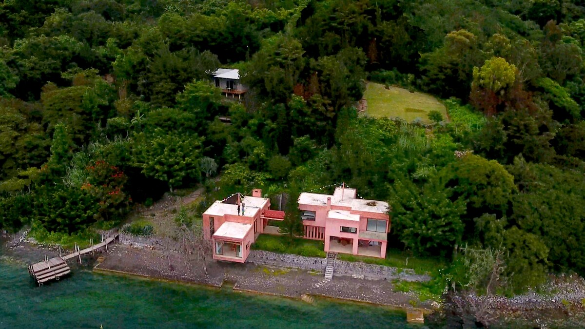 An aerial view of a modern pink house situated along a lakeshore, surrounded by lush greenery. A private dock extends into the water, providing easy access. The property encompasses landscaped gardens and neighboring structures, blending harmoniously with the natural environment.