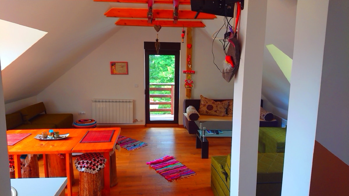The spacious living area features natural wood accents and a comfortable seating arrangement, including a sofa and armchairs. A wooden table is centrally placed, surrounded by colorful textiles. Large windows allow for natural light, while views of greenery enhance the relaxed ambiance.
