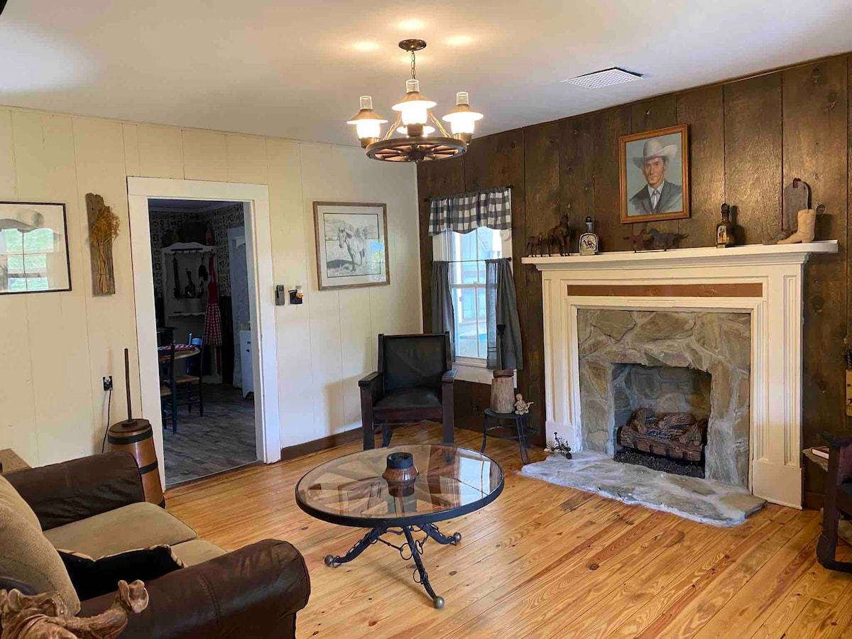 A cozy living area features a stone fireplace, framed artwork, and wooden walls, with natural light coming through a window. A round glass coffee table sits on a wooden floor, surrounded by a sofa and two armchairs, creating a welcoming space.