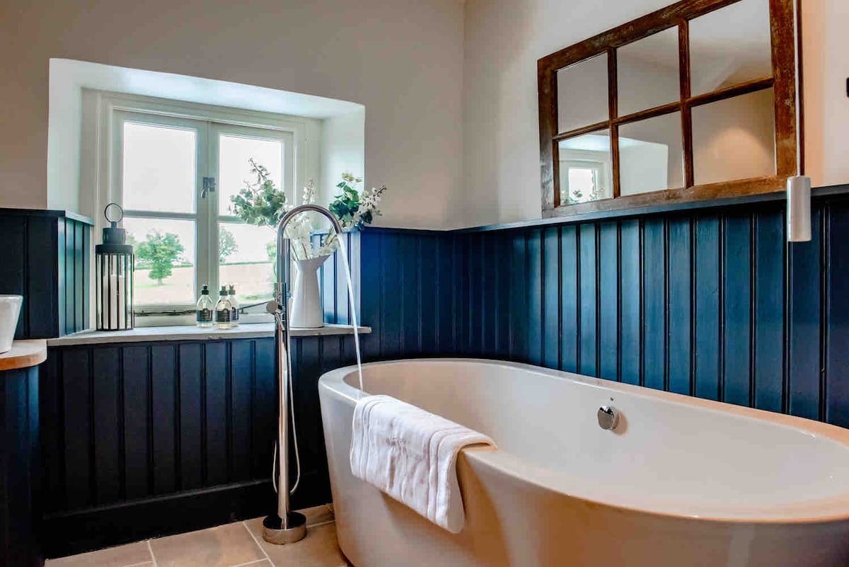 A spacious bathroom is highlighted by a freestanding bathtub positioned beneath a window. Dark blue paneling contrasts with light-colored tiles, while botanical accents are visible on the windowsill. A modern faucet stands next to the tub, completing the elegant look of the space.