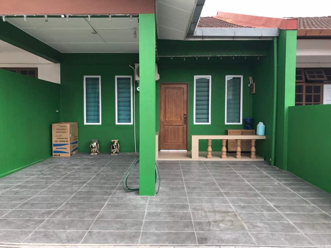 An exterior view of the guesthouse is presented, featuring vibrant green walls and a tiled floor. Two windows with lightweight curtains frame the inviting entrance, which is complemented by a wooden door. A few seating elements and storage boxes are arranged along the side.