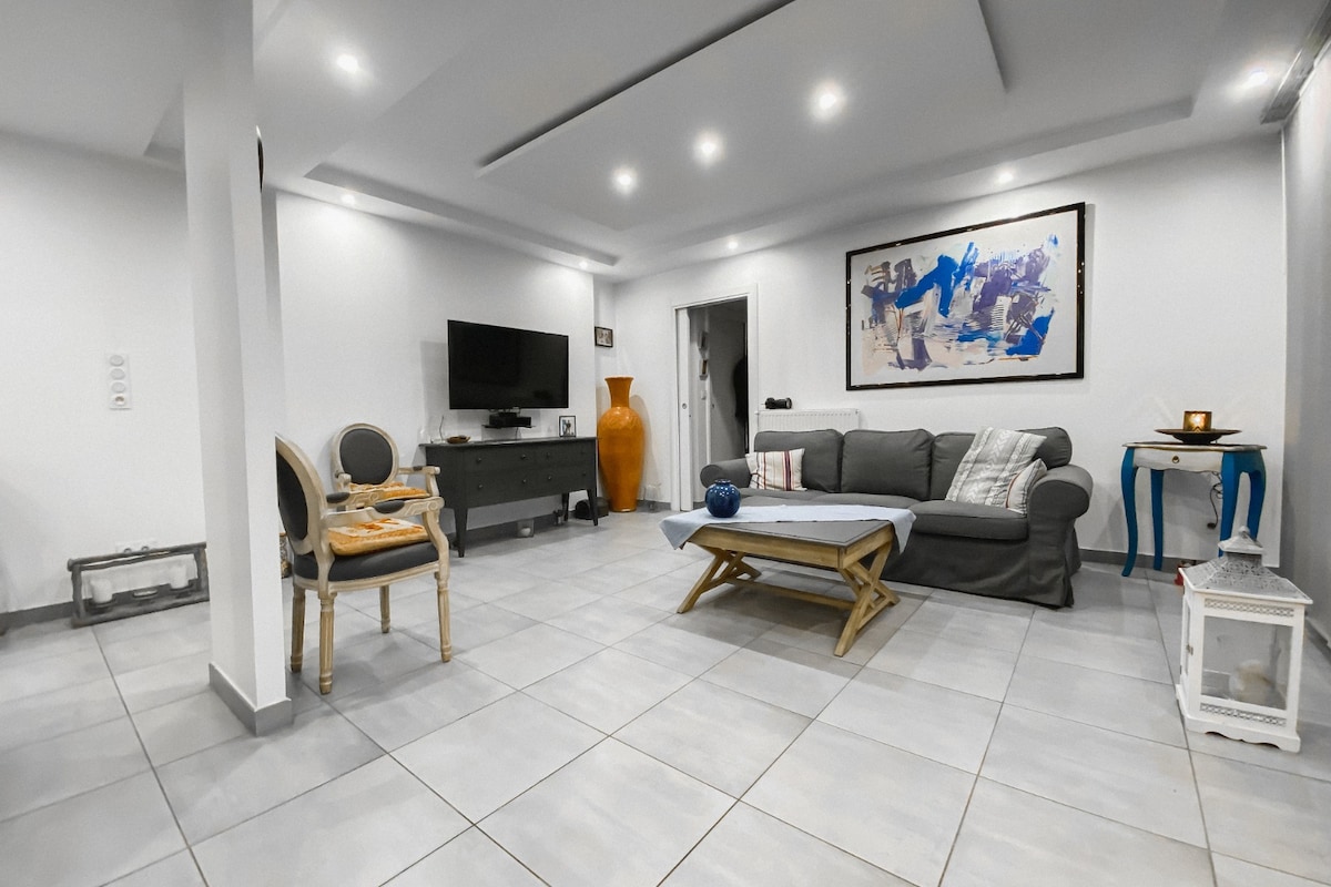 A well-lit living area features modern furnishings, including a grey sofa and a light wooden coffee table. A large piece of artwork hangs on the wall, while a decorative vase and a small table are positioned in the corners. The tiled floor adds a clean and elegant touch.