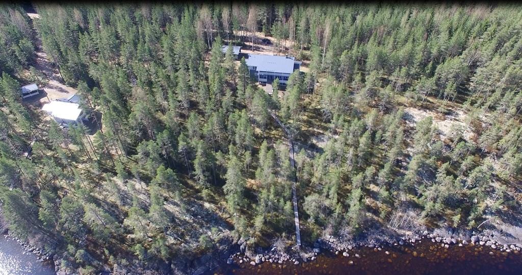 Aerial view of lush greenery surrounding a modern building situated near the edge of a lake. A clear path extends from the structure towards the water's edge, where rocks border the shoreline.