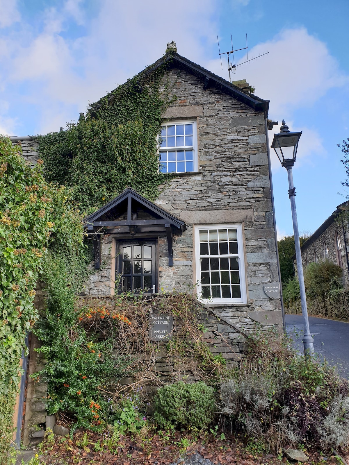 The exterior of a charming stone cottage is featured, surrounded by lush greenery. Tall windows provide a glimpse of the interior. A lantern stands nearby, adding to the cottage's quaint character. The building reflects traditional architecture within a serene environment.