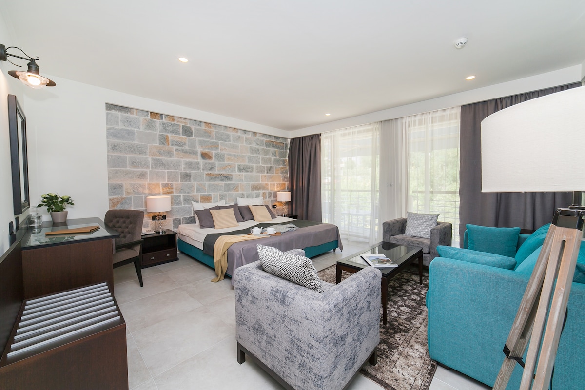 A spacious junior suite features a mix of modern and natural design elements. A large bed is complemented by a lounge area with two armchairs and a sofa. Floor-to-ceiling windows allow natural light, while a stone accent wall adds a unique touch.