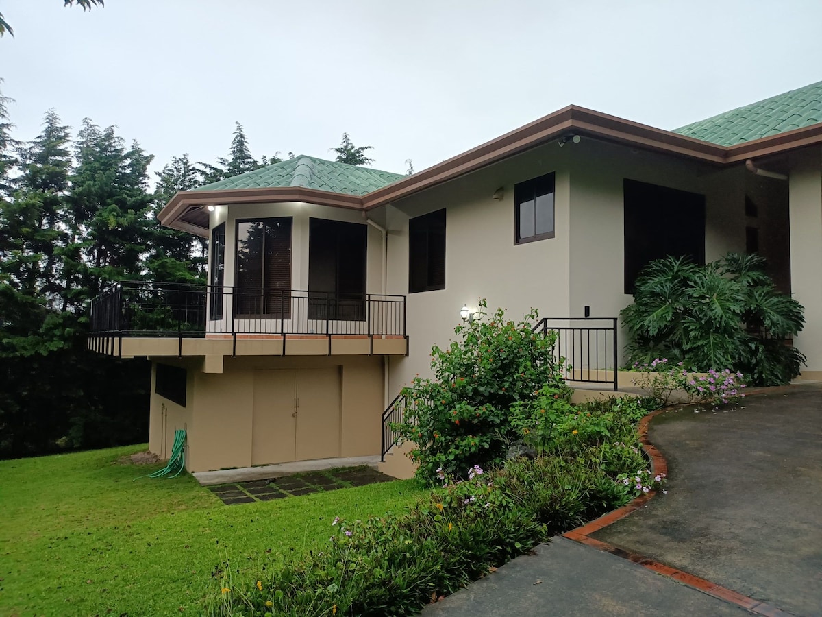 A modern house featuring a green metal roof and large windows, nestled among lush greenery. A gradual driveway leads up to the entrance, which is complemented by neatly trimmed grass and flowering plants. A balcony extends from the second level, providing a spacious outdoor area.