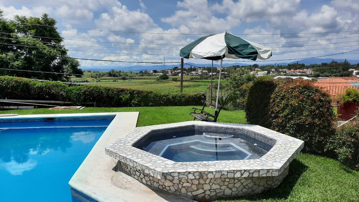 A spacious outdoor area features a rectangular pool alongside a circular jacuzzi. A shaded seating structure is positioned nearby, overlooking lush green grass and vibrant gardens. The surrounding hills and distant landscape provide a scenic backdrop under a partly cloudy sky.
