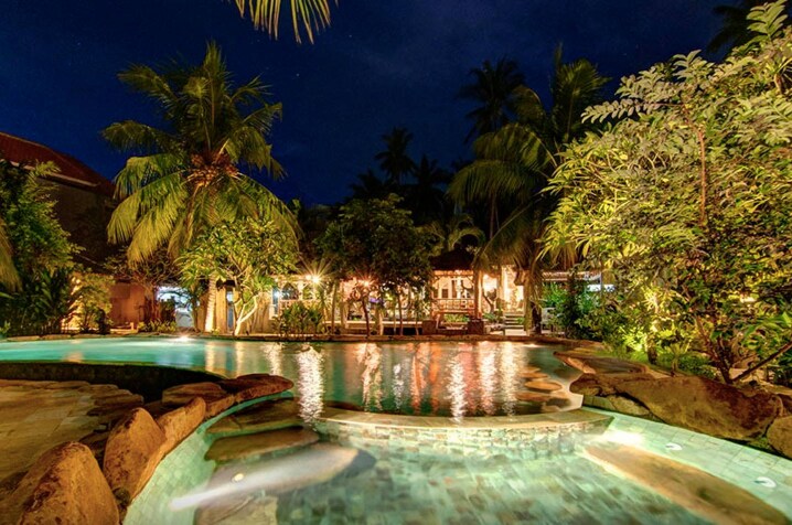 A tranquil swimming pool is surrounded by lush palm trees and tropical plants, reflecting soft lighting from nearby structures. The water's surface glimmers under a night sky, creating an atmosphere of calm. Steps lead into the pool, enhancing accessibility.