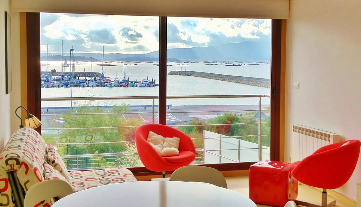 A spacious living area is shown, featuring large floor-to-ceiling windows that offer a panoramic view of the marina and waterfront. Bright red plush chairs complement a round white table, with natural light flooding the space. The tranquil water reflects the cloudy sky, enhancing the serene atmosphere.