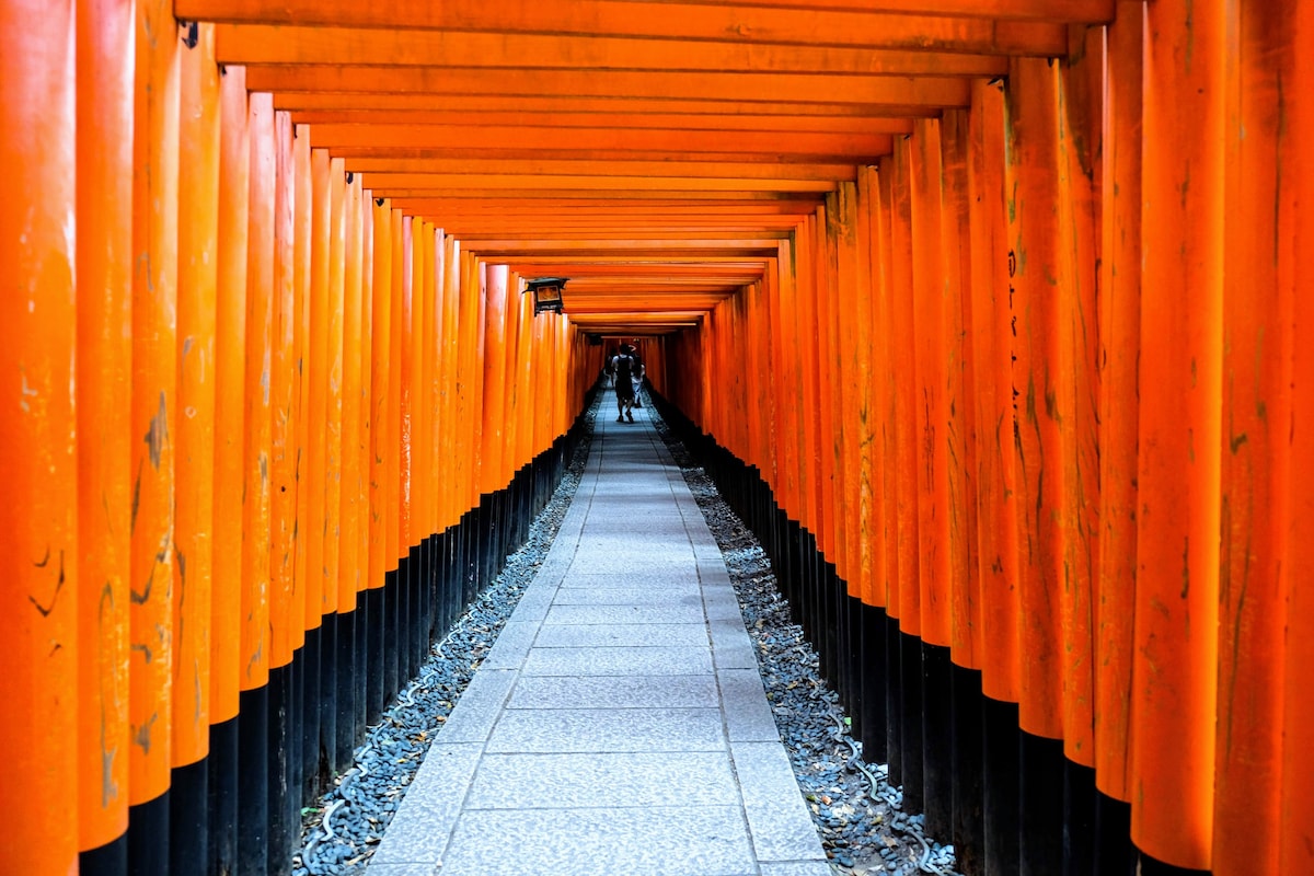 A corridor lined with vibrant orange torii gates creates a striking pathway. The stone-paved walkway is flanked by uneven gravel, guiding visitors through this iconic site. Dim lighting from overhead lanterns adds a serene ambiance to the space.
