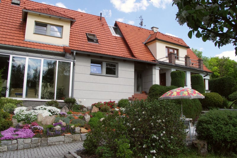 The exterior of a family-style guesthouse is showcased, featuring a red-tiled roof and multiple windows. A well-maintained garden, adorned with colorful flowers and shrubs, is visible in the foreground. A patio umbrella provides shade near a seating area, enhancing the inviting outdoor space.