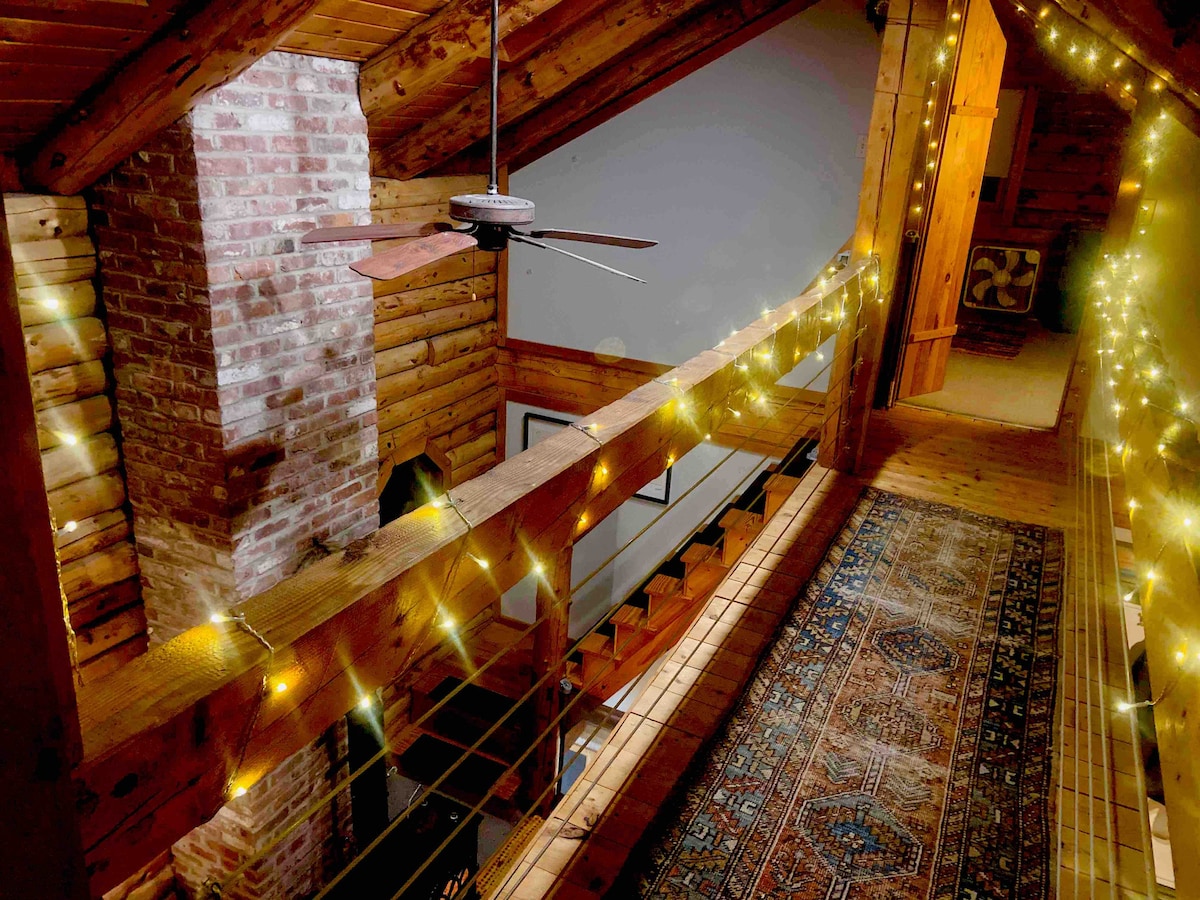 A warmly lit loft space features a wooden railing adorned with string lights. A rustic ceiling fan hangs from the exposed beams, and a patterned area rug lies along the wooden floor. The brick wall adds a touch of texture to the inviting atmosphere.