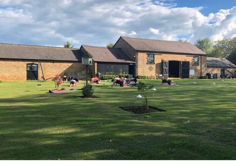 A spacious outdoor area is shown, featuring a grassy lawn in front of a large barn structure. Several individuals practice yoga on mats, while the barn's brick facade and large doors add rustic charm. Trees provide shade, and the sky is partially cloudy.