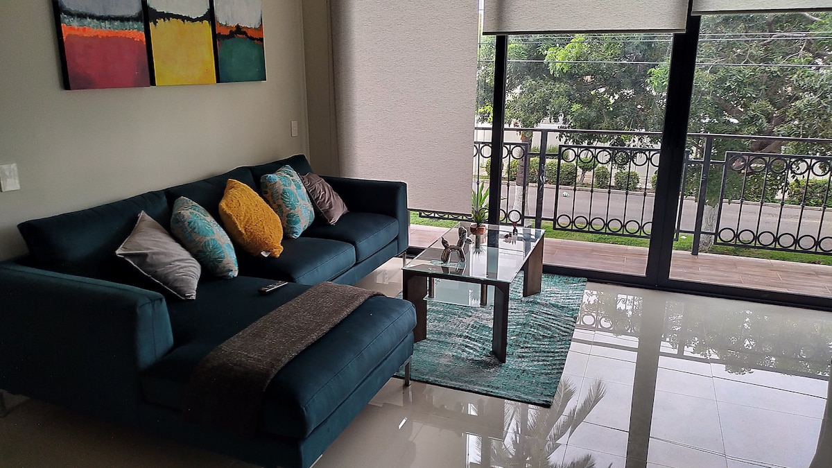 A modern living area features a comfortable sectional sofa adorned with colorful decorative pillows. A glass coffee table rests on a soft area rug, with natural light filtering through large windows that open to a balcony overlooking lush greenery.